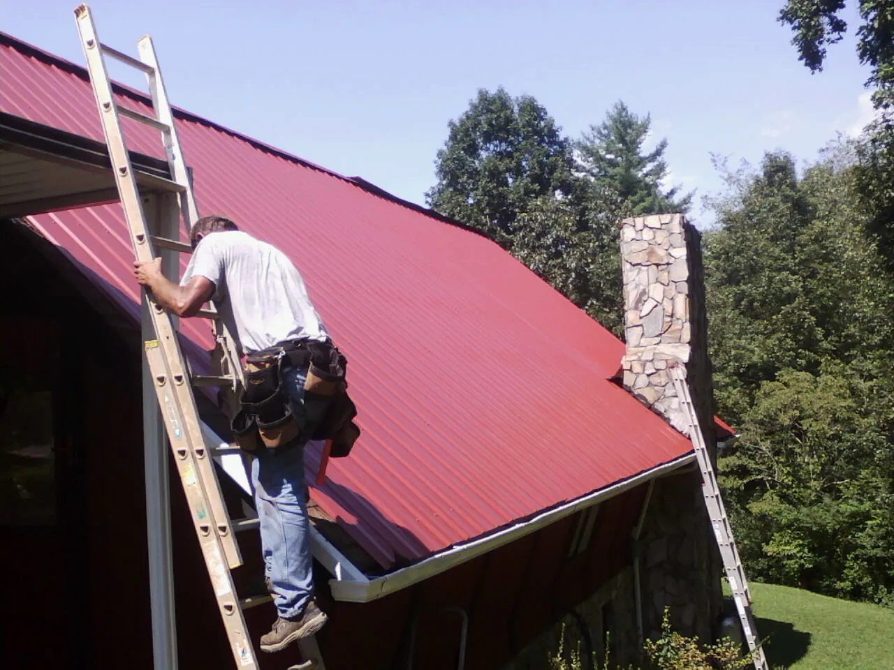 Roofer on a ladder beside a red metal roof and stone chimney — Soffit & Fascia Repair by Roofing Brothers in Loma